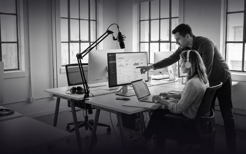 Photo en noir et blanc : dans un studio de digital learning, une professeure de français casque sur les oreilles relit une copie annotée sur son ordinateur pendant qu'un chef de produit lui montre un tableau de bord à l'écran.