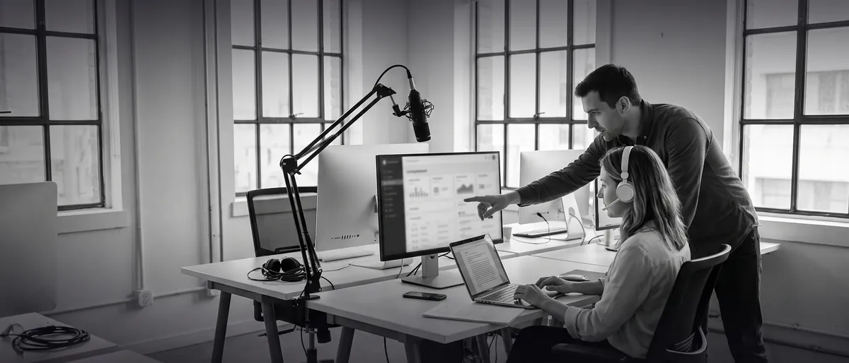 Photo en noir et blanc : dans un studio de digital learning, une professeure de français casque sur les oreilles relit une copie annotée sur son ordinateur pendant qu'un chef de produit lui montre un tableau de bord à l'écran.